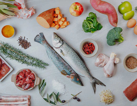 Various ingredients laid out, including a whole fish, vegetables, herbs, spices, and meats on a pale background.
