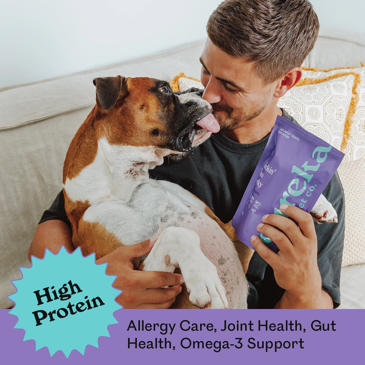 Man cuddling a happy Boxer dog on a couch while holding a purple bag of Eureka Pet Co. Rockin Roo Jerky — high protein, allergy care, joint health, gut health and omega-3 support
