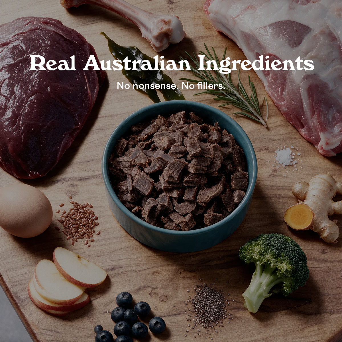 Blue bowl of Eureka Pet Co air-dried dog food surrounded by raw ingredients on a wooden board, including kangaroo meat, a lamb bone, kelp, rosemary, chicory root, egg, flaxseed, apple, blueberries, chia seeds and broccoli. Text reads Real Australian Ingredients, no nonsense, no fillers.