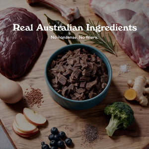 Blue bowl of Eureka Pet Co air-dried dog food surrounded by raw ingredients on a wooden board, including kangaroo meat, a lamb bone, kelp, rosemary, chicory root, egg, flaxseed, apple, blueberries, chia seeds and broccoli. Text reads Real Australian Ingredients, no nonsense, no fillers.