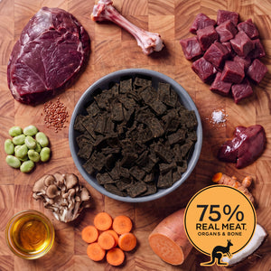 Blue-grey bowl of Eureka Pet Co air-dried dog food surrounded by raw ingredients on a wooden board, including a bone, kangaroo meat, broad beans, coconut, flaxseed, sweet potato, turmeric and carrots. Badge reads 75% real meat, organs and bone.