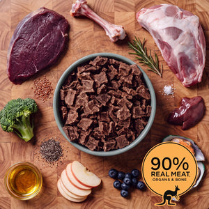 Grey bowl of Eureka Pet Co air-dried dog food surrounded by raw ingredients on a wooden board, including kangaroo meat, a bone, lamb shoulder, rosemary, ginger, flaxseed, apple, blueberries and chia seeds. Badge reads 90% real meat, organs and bone.