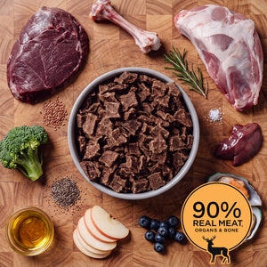 Grey bowl of Eureka Pet Co air-dried dog food surrounded by raw ingredients on a wooden board, including venison, a bone, lamb shoulder, rosemary, ginger, flaxseed, apple, blueberries and chia seeds. Badge reads 90% real meat, organs and bone.
