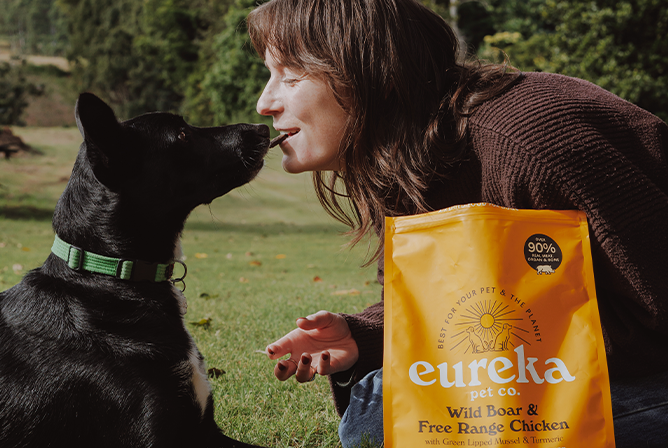 Woman bonding with a black dog in a park with a bag of Eureka Wild Boar & Free Range Chicken dog food.