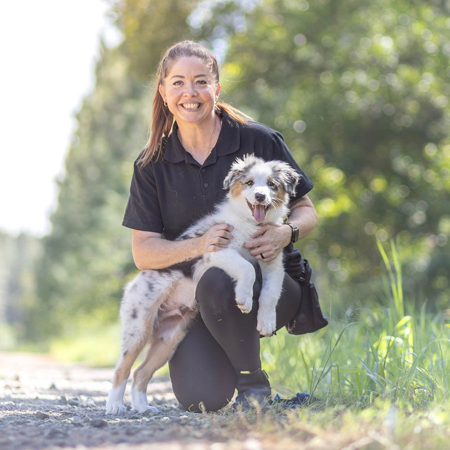 Jazz, The Everyday Dog Trainer crouching in the park with a cute Aussie Shepard puppy in her arms.